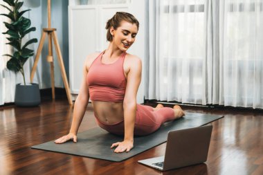 Spor kıyafetleri içinde esnek ve becerikli bir kadın evde egzersiz paspası üzerinde meditasyon pozisyonunda yoga yapıyor. Huzurlu bir zihin ve sükunetle evde yoga eğitimi..
