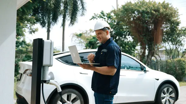 Qualified technician working on home EV charging station installation, making troubleshooting and configuration setup on charging system with laptop for EV at home. Synchronos