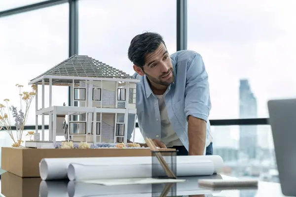 Professional caucasian architect engineer working with house model turning head at laptop on table with project plan, architectural model. Young businessman thinking about investment. Tracery.