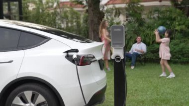 Mutlu ve neşeli bir ailenin arka planında ev şarj istasyonundan elektrikli araç şarj ediliyor. EV arabası daha iyi bir gelecek için alternatif ve sürdürülebilir enerji kullanıyor.