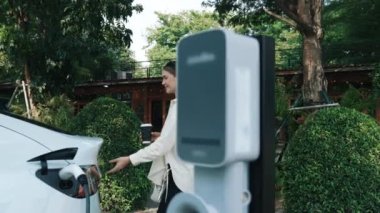 Yavaş çekimde çalışan kadın ve EV elektrikli araba ile sürdürülebilir şehir ulaşımı ilkbahar bahçesindeki açık kafede şarj oluyor, yeşil şehir sürdürülebilirliği ve çevre dostu EV otomobil. Yararlı