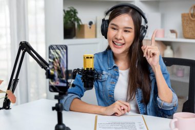 Asyalı sunucu canlı yayında canlı yayında canlı yayında sosyal medyada kulaklık takarak konuşuyor. Dinleyicileri modern stüdyoda koçluk yaparak selamlıyor. Taktik.