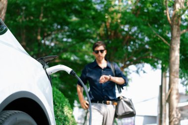 Genç adam baharda açık yeşil şehir parkındaki şarj istasyonundan elektrikli araba aküsünü şarj ediyor. Sürdürülebilir çevre dostu kentsel seyahat tarzı için şarj edilebilir EV araba. Yararlı