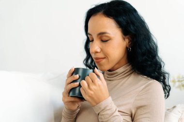 Evdeki kanepede kahve içen mutlu kadın dinlenmek ve dinlenmek için çok önemli. Elinde fincanla genç bir Afrikalı Amerikalı kadının portresi..