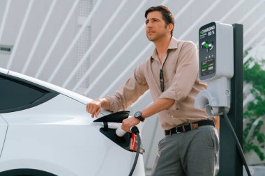 Genç adam, şehrin ticari otoparkındaki şarj istasyonundan elektrikli araba aküsünü şarj etmek için EV şarj aleti koymuş. Sürdürülebilir çevre dostu şehir yolculuğu için şarj edilebilir EV araba. Yararlı