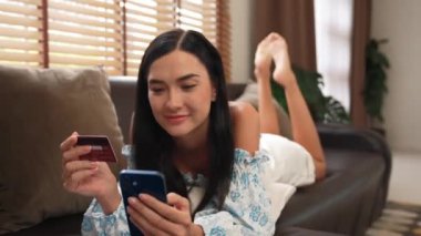 Kanepede yatan genç bir kadın internet bankacılığı için kredi kartıyla dizüstü bilgisayar kullanıyor. Evdeki online ödeme kapısından alışveriş yapıyor. Modern ve uygun online satın alma. Blithe