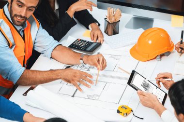 Mimari proje üzerinde birlikte çalışan çeşitli inşaat mühendisi ve müvekkil grupları, inşaat planını gözden geçirip toplantı masasında plan oluşturuyorlar. Tedbirli