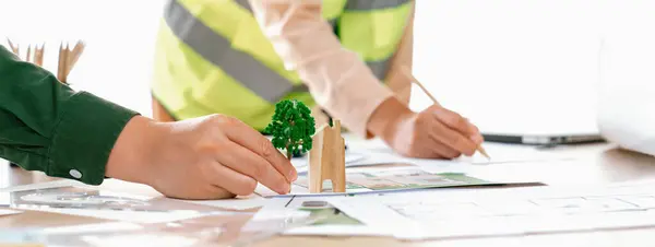 Professional architect and engineer collaborate to design eco-friendly house on meeting table with blueprint, wooden house block and tree model scatter around. Closeup. Delineation.