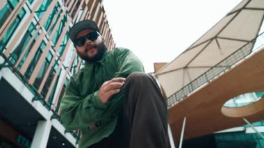 Hip hop tarzında düşük açılı hipster pozu alışveriş merkezi veya bina manzarası ile çevrilidir. Kameraya bakarken Asyalı hip hop koreografı break dans yapıyor. Açık hava sporu 2024 hiphop..