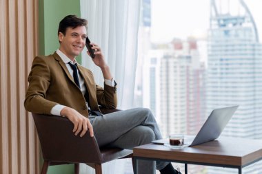 Gökdelen manzaralı pencerenin yanında otururken yakışıklı iş adamının müdürle telefon görüşmesi yaptığı yakın plan. Yönetici, telefon ve dizüstü bilgisayarla konuşuyor. Önüne bak. Süslü.