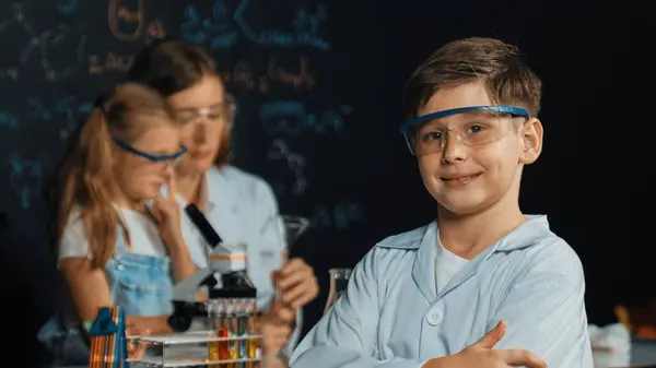 Smart boy pour chemical liquid in beaker while teacher giving advice at STEM science class. Student mixing colored solution while standing at board written chemistry theory at laboratory. Erudition.