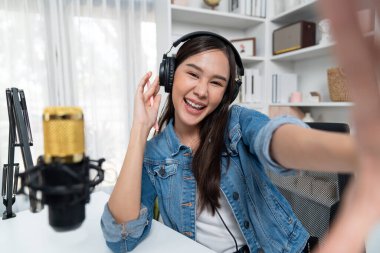 Sunucu kanalı genç ve güzel Asyalı kız akıllı telefonuyla selfie çekiyor. Modern stüdyo ofis konseptinde kulaklık takıyor. Sahne arkası yayıncısı sosyal medyada canlı yayın yapıyor. Taktik.