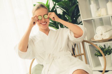 Elinde taze salatalık dilimleri tutan ve bornoz giyen bir kadın tatil köyünde ya da otelde lüks yüz derisi kaplıcalarının tadını çıkarıyor. Yüz ve güzellik tedavisi için cilt bakımı. Sessiz.