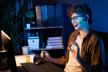 Genç yaratıcı iş adamı, bilgisayar ekranında dinleyicilerle iletişim kuruyor ya da takım üyesiyle görüşüyor. Neon Light Modern Ev Ofisi 'nde kulaklık takıyor. Gece vakti çalışma kavramı. Toplayıcı.