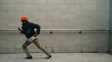 Asyalı akıllı hipster, kulaklık ve şık kıyafetler giyip hip hop şarkıları dinlerken gri arka planlı enerjik break dans gösterisi yapıyor. Açık hava sporu 2024. Hip hop hareketi. Yumuşak bir şekilde..