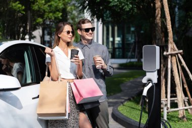 Genç çift yazın yeşil sürdürülebilir şehir bahçesinde EV elektrikli araba ile seyahat ederken, yeşil temiz yeniden şarj edilebilir elektrikli araç iç organları ile kentsel sürdürülebilirlik yaşam tarzını gösteriyor.