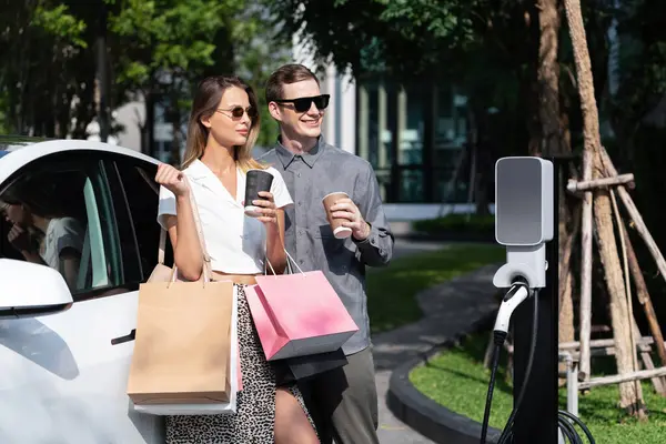 Genç çift yazın yeşil sürdürülebilir şehir bahçesinde EV elektrikli araba ile seyahat ederken, yeşil temiz yeniden şarj edilebilir elektrikli araç iç organları ile kentsel sürdürülebilirlik yaşam tarzını gösteriyor.