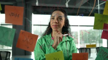 Pazarlama fikrini düşünen yetenekli, genç ve güzel İspanyol proje yöneticisinin portresi. Ofisteki cam tahtada yapışkan notlar kullanarak iş planı yapan profesyonel bir iş kadını. Yönlendirici.