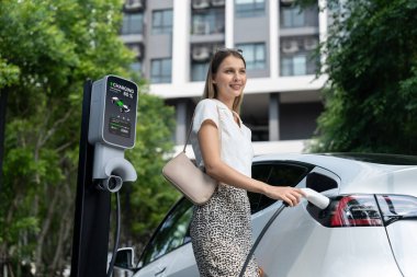 Genç bir kadın yazın yeşil sürdürülebilir şehir bahçesinde EV elektrikli arabasıyla seyahat ediyor. Kentsel sürdürülebilirlik yaşam tarzı yeşil temiz yeniden şarj edilebilir enerji elektrikli BEV araç iç organları