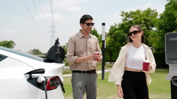 两对年轻夫妇在与电网塔电力工业设施相连的充电站为电动汽车充电 为生态友好型汽车的使用提供了动力 — 图库视频影像