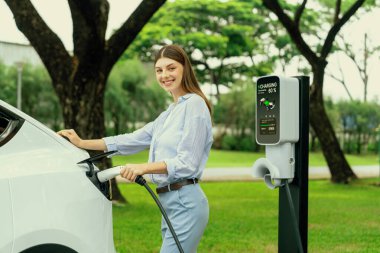 Açık yeşil şehir parkı manzaralı EV şarj istasyonundan genç kadın EV elektrikli araçları şarj ediyor. Çevre dostu şehir ulaşımı ve çevre dostu EV araba seyahati. Yükselt