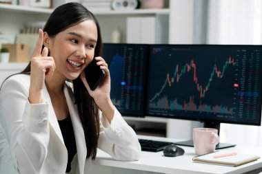 Genç ve güzel Asyalı iş kadını, modern ofisteki artan borsa grafiğini analiz etmek için bilgisayar ekranından brokerla telefonda konuşuyor ve zenginliğe yüksek miktarda yatırım yapıyor. Taktik.