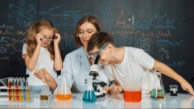 Öğretmen tavsiye verirken, beyaz çocuk mikroskop altında bakıyor. Profesyonel eğitmen labaratuar kıyafeti giymiş, renkli solüsyonla dolu deney kabıyla sofrada çeşitli öğrencileri arıyor. Etkinlik.