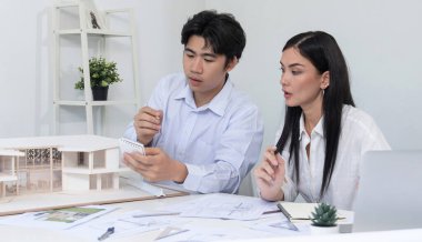 Profesyonel Asyalı erkek mimar ev modelinin uzunluğunu ölçmek için cetvel kullanırken, beyaz tenli genç meslektaşı ev modeliyle toplantı masasındaki verileri analiz etmek için dizüstü bilgisayar kullanıyor. Tertemiz..