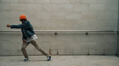 Asyalı akıllı hipster, kulaklık ve şık kıyafetler giyip hip hop şarkıları dinlerken gri arka planlı enerjik break dans gösterisi yapıyor. Açık hava sporu 2024. Hip hop hareketi. Yumuşak bir şekilde..