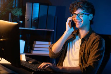 Genç iş adamı akıllı telefondan müşteriyi ya da iş arkadaşını arıyor, neon modern ofisteki yaratıcı sunum surround çalışma masasının bilgisayar ekranına bakıyor. Yeni proje kavramı. Toplayıcı.