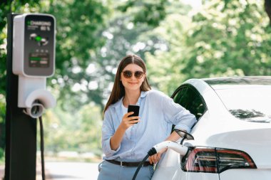 Ulusal Park veya Yaz Ormanı 'ndaki tatil gezisi sırasında EV şarj istasyonundan elektrik aküsü almak için akıllı telefon internet bankacılık uygulaması kullanan genç bir kadın. Yükselt
