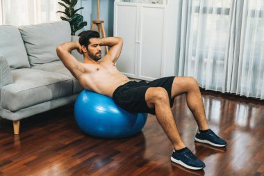 Sporcu ve atletik bir adam evde vücut egzersizi yaparken spor topunun üzerinde oturarak formda fiziki ve sağlıklı spor yaşam tarzına uygun bir yaşam sürüyor. Gaiety ev egzersizi antrenman konsepti.