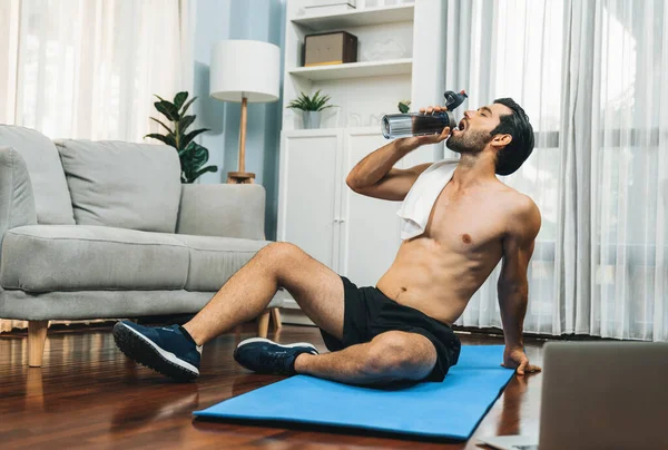 Athletic and sporty man drinking water on fitness mat after finishing home body workout exercise session for fit physique and healthy sport lifestyle at home. Gaiety home exercise workout training.