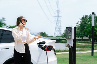EV araba aküsünü şarj eden genç bir kadın elektrik şebekesine bağlı elektrik santralinde çevre dostu araç kullanımı için elektrik endüstrisi olarak telefonla konuşurken. Yararlı