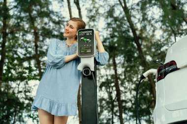 Yeşil kırsal kesimde tatil gezisi. Güzel genç bir kadın alternatif enerjiyle elektrikli aracı şarj ediyor. Çevre dostu seyahat akıllı EV araba. Kalıcı