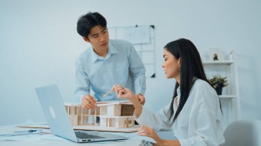 Profesyonel Asyalı erkek mimar ev modelinin uzunluğunu ölçmek için cetvel kullanırken, beyaz tenli genç meslektaşı ev modeliyle toplantı masasındaki verileri analiz etmek için dizüstü bilgisayar kullanıyor. Tertemiz..