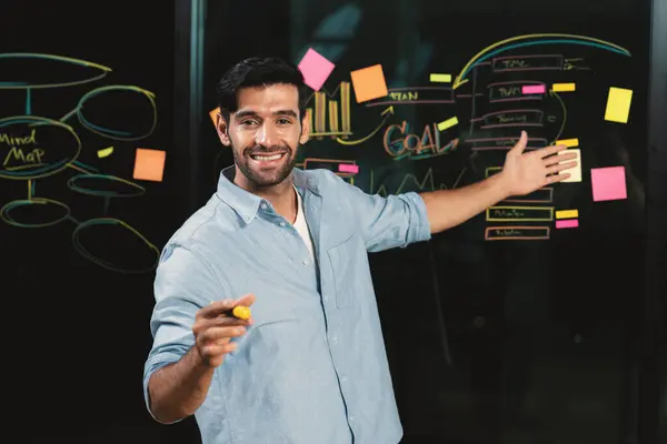 Professional businessman pointing at marketing statistic while looking at camera. Smiling caucasian manager presenting about business plan with confident at glass wall by graph, mind map. Tracery.