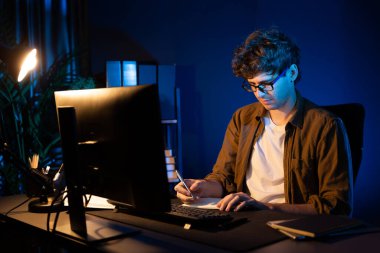 Genç işadamı, Neon Light Modern Ofis 'teki yeni proje surround çalışma masası için kağıt üzerinde tasarım düşünüyor. E-posta ile hediye göndermeden önce not defterine yeni bir iş düşünüyorum. Toplayıcı.