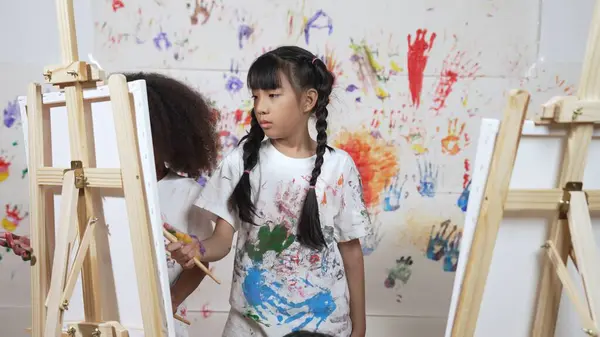 Playful student painted or draw canvas at stained wall with diverse friends in art lesson. Group of happy multicultural children working or create artwork at messy room. Creative activity. Erudition.