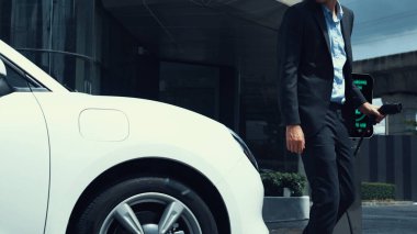 İş adamı elektrikli arabasını şehir merkezindeki şarj istasyonundan ya da otoparktan şarj ediyor. Şehir yaşam tarzında alternatif temiz enerji kullanan çevre dostu şarj edilebilir araba. İnceleyin.