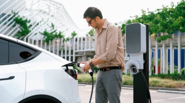 Genç adam, şehrin ticari otoparkındaki şarj istasyonundan elektrikli araba aküsünü şarj etmek için EV şarj aleti koymuş. Sürdürülebilir çevre dostu şehir yolculuğu için şarj edilebilir EV araba. Yararlı