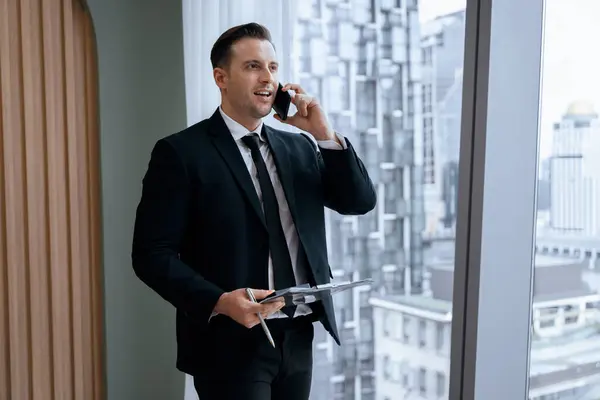Portrait of professional businessman stand near window with skyscraper while phone calling and holding laptop. Busy executive manager holding laptop while multitasking working. Closeup. Ornamented.