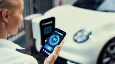 Akıllı telefon akü durumu arayüzünü elinde tutan iş kadını akıllı EV mobil uygulama ile otomobil parkındaki şarj istasyonundan elektrik şarj eden çevre dostu EV araba. İnceleyin