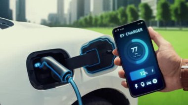 İşadamı çevre yeşil şehir parkındaki şarj istasyonundan şarj olurken akıllı telefon ekranından EV batarya durum mobil uygulamasını kontrol ediyor. Elektrikli arabanın gelecekteki enerji sürdürülebilirliği. İnceleyin