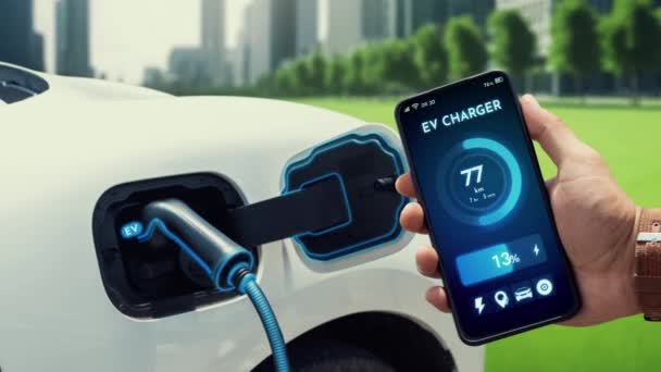 ビジネスマンは エコシティパーク内の充電ステーションから充電しながら スマートフォン画面上でEvバッテリーのステータスモバイルアプリケーションを確認します 電気自動車の未来的なエネルギー持続可能性 Peruse — ストック動画