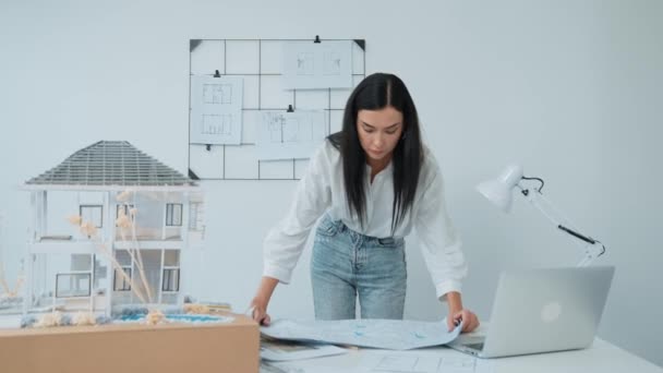 若い美しい白人建築家は テーブルに青写真を置き 家のモデルと比較しながら 現代のオフィスでホースモデルの構造を注意深く検査し 青写真をストライプ インマキュライト — ストック動画