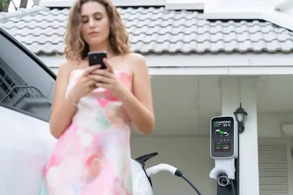以家庭电动车充电站为重点 模糊背景下的现代环保妇女使用智能手机 住宅概念的电动车技术应用 同步操作 — 图库照片