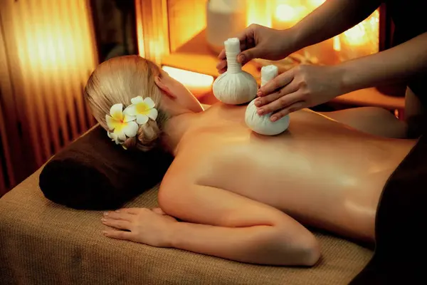 Hot herbal ball spa massage body treatment, masseur gently compresses herb bag on woman body. Tranquil and serenity of aromatherapy recreation in warm lighting of candles at spa salon. Quiescent