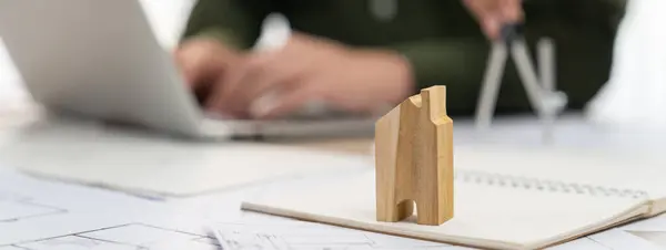 Wooden block was placed on table with blueprint and architectural document scatter around while professional architecture working on laptop behind. Blurring background. Cropped image. Delineation.