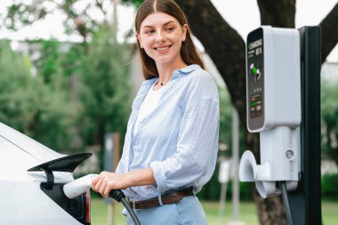 Açık yeşil şehir parkı manzaralı EV şarj istasyonundan genç kadın EV elektrikli araçları şarj ediyor. Çevre dostu şehir ulaşımı ve çevre dostu EV araba seyahati. Yükselt